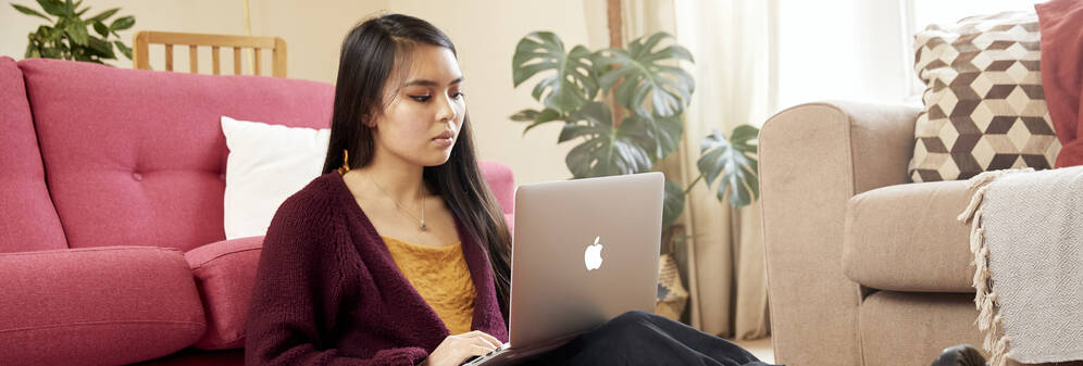Female student sitting on the floor leaning on a sofa with a laptop on her knees. She is looking at the laptop and applying for student accommodation on the University of Bristol